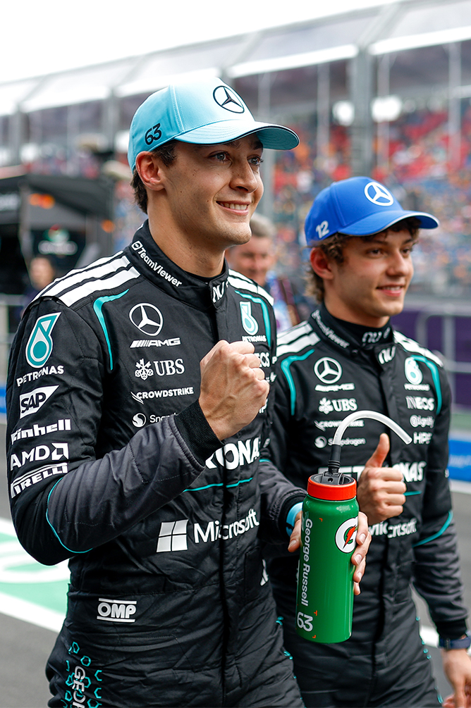Two Formula 1 drivers with helmets and drink bottles at the racetrack.