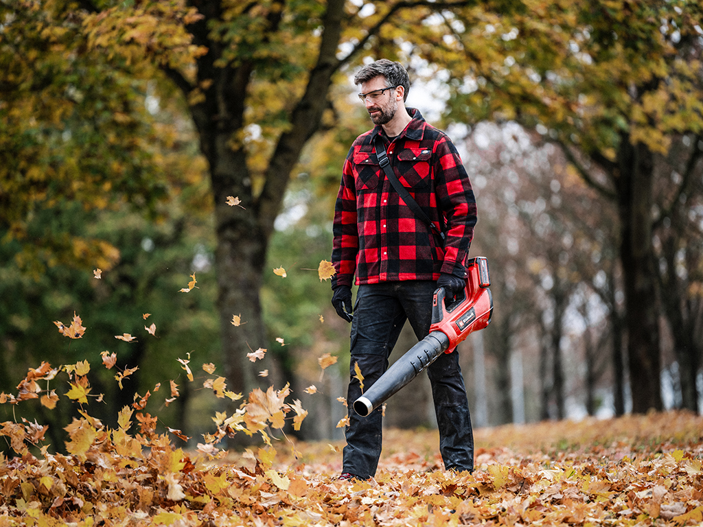 The picture shows a man wearing a red and black checked jacket using an Einhell cordless leaf blower to blow leaves into the air and away.
